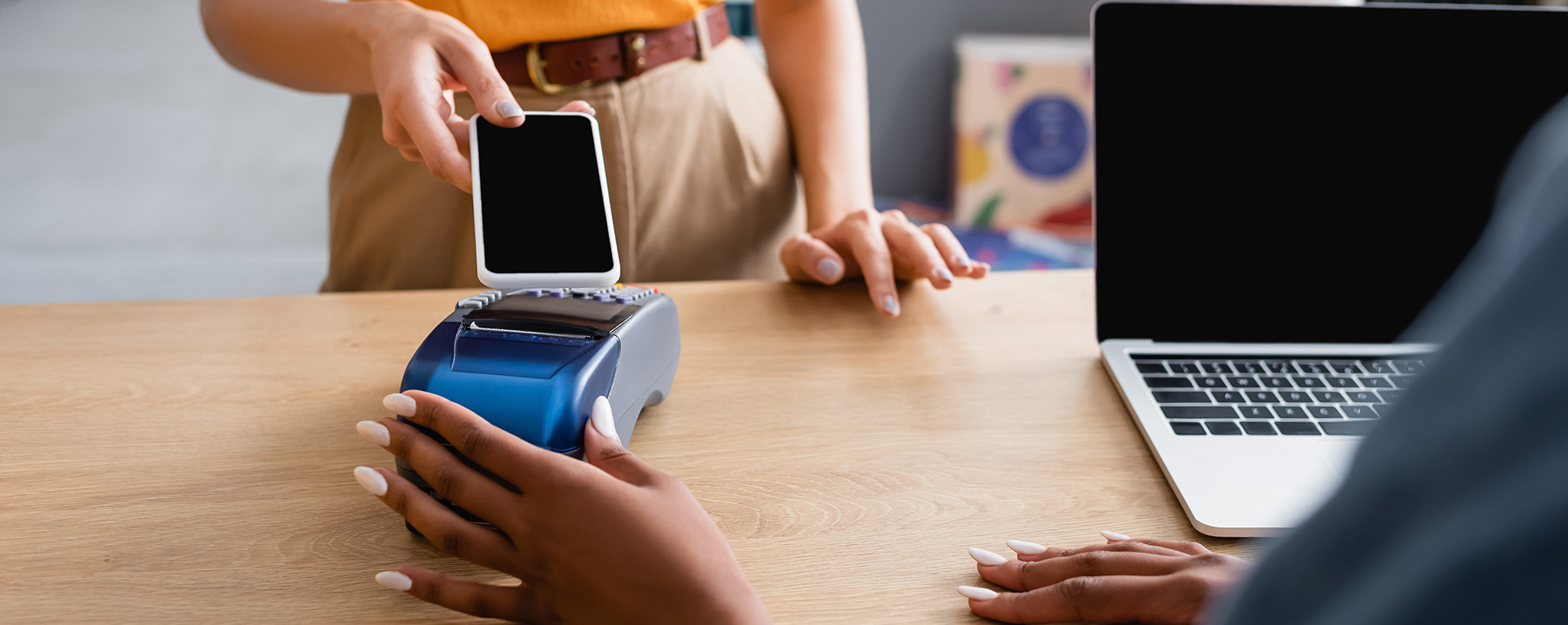cropped view of woman paying with smartphone near laptop and african american saleswoman in boutique